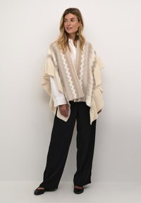 Beige strikket poncho med zigzag mønster og tekstureret kant, parret med en hvid skjorte med krave og sorte breddebenede bukser. Flade sorte sko.