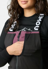 Schwarzer isolierter Overall mit lila Akzenten, Reißverschlusstasche, verstellbaren Trägern mit "mou"-Logo, getragen über einem schwarzen Langarmshirt.