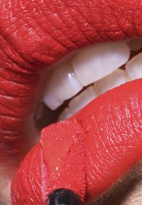 Rød leppestift på glatte, teksturerte lepper med en blank finish, som fremhever hvite tenner som delvis er synlige mellom leppene.