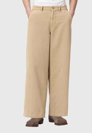 Person trägt beige, weit geschnittene Hosen, hat die Hände in den Taschen und trägt braune Wildlederstiefel, steht vor einem grauen Hintergrund.