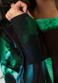 Nahaufnahme einer Hand, die den Bund eines langärmeligen, grün-schwarz gemusterten Kleidungsstücks mit einem türkisfarbenen Banddetail anpasst.