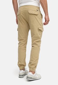 Pantalones cargo caqui con piernas ajustadas, puños elásticos y múltiples bolsillos. Llevados con zapatillas gris y blancas. Presentan una textura suave.