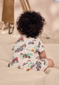 Vêtement pour bébé en tissu blanc avec des motifs de tracteurs colorés, à manches et jambes courtes. Texture douce, conçu pour le confort, vu de l'arrière.