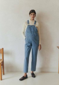 Svetlo modre denim overall hlače imajo široke naramnice, sprednje žepke in ohlajen kroj. So v kombinaciji s krem puloverjem in temnimi ravnimi čevlji.