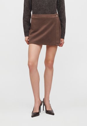 Femme portant une mini-jupe marron avec un petit détail métallique à la taille, associée à un pull foncé et des chaussures à talons noirs.