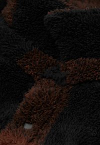 Svart och brunt fleece tyg med en texturerad, fluffig yta. Har en synlig knappdetalj och kontrasterande färgpatchar i designen.