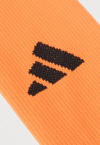 Πορτοκαλί κάλτσα με υφασμένο μοτίβο, που διαθέτει ένα μαύρο λογότυπο αποτελούμενο από τρεις διαγώνιες ρίγες κοντά στην κορυφή.