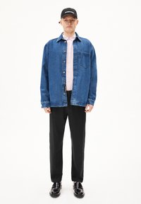 Blauwe spijkerjacket met een borstzak, gelaagd over een lichtroze shirt, gecombineerd met zwarte broeken en zwarte schoenen. Zwarte pet met logo.