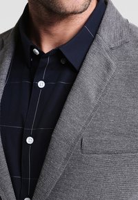 Harmaapintainen bleiseri lovikauluksin tummansinisen, ruudukkomallisen nappipaidan päällä. Nappien ja taskun yksityiskohdat näkyvissä.