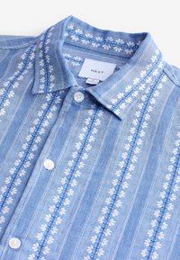 Chemise bleu clair à col avec motifs floraux blancs, rayures verticales et deux boutons. Étiquettes visibles dans la zone du col.