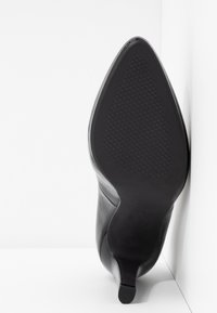 Chaussure à talon haut en cuir noir à bout pointu reposant semelle vers le haut contre un mur blanc en coin, montrant le motif texturé de la semelle.