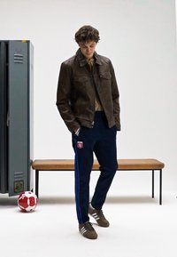 Brun skinnjakke, beige skjorte, marineblå joggebukser med sidestreker og brune joggesko. Fotball i nærheten, grå skap i bakgrunnen.