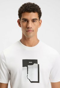 Hvid kortærmet t-shirt med en sort og hvid grafisk tryk med et design af revnet papir og "BOSS" tekst øverst. Glat bomuldsstof.