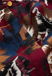 Flerfärgad tyg med ett blommigt och abstrakt mönster i röd, blå, brun och svart. Slät textur med synlig sömnad och en gyllene knapp.