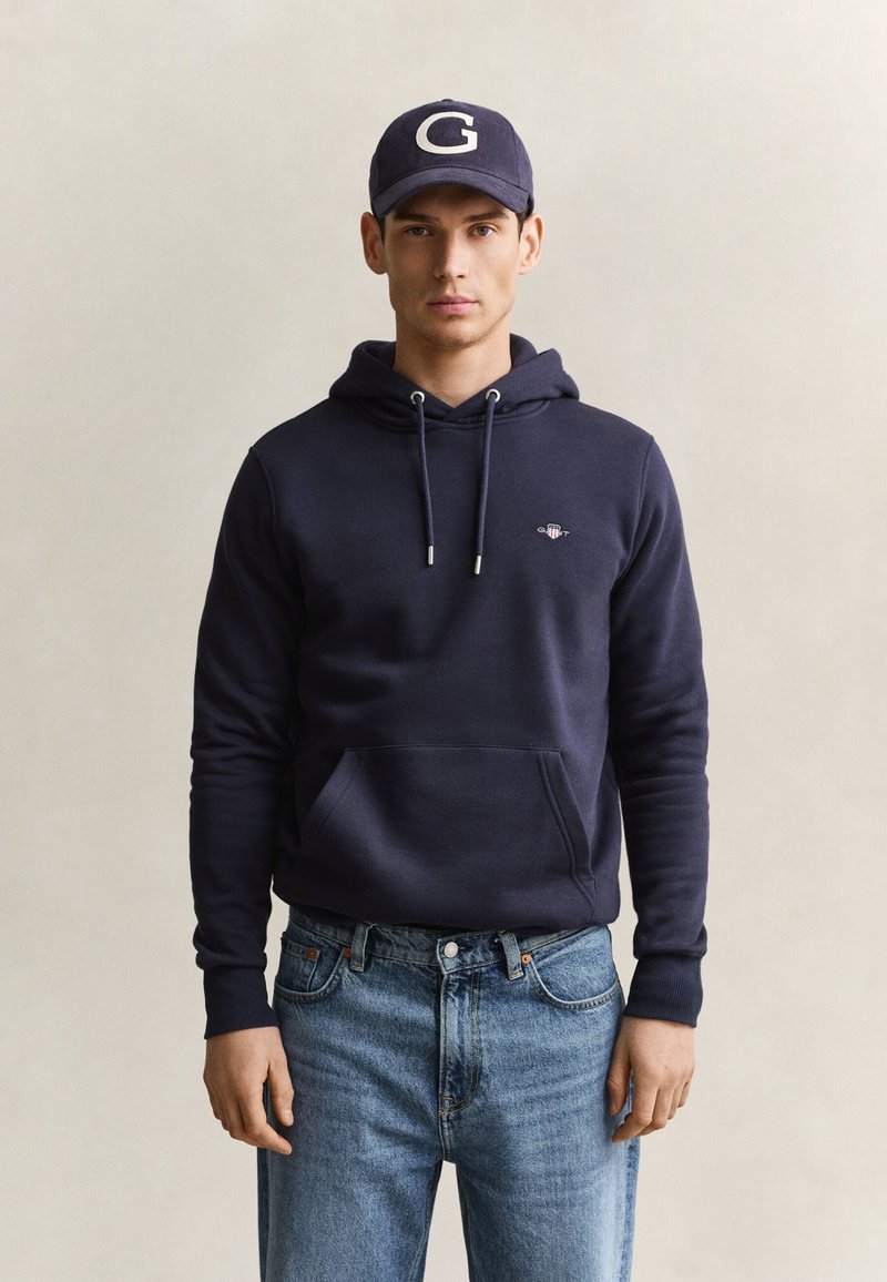 Jeune homme portant un sweat à capuche bleu marine avec cordons, jean bleu et casquette bleu marine avec la lettre blanche G, debout devant un fond clair uni.