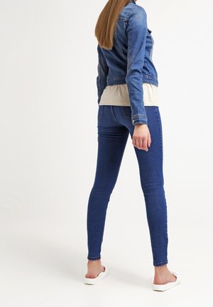 Femme debout de dos portant une veste en jean bleue, un jean skinny bleu, un haut beige et des chaussures blanches slip-on sur fond blanc.