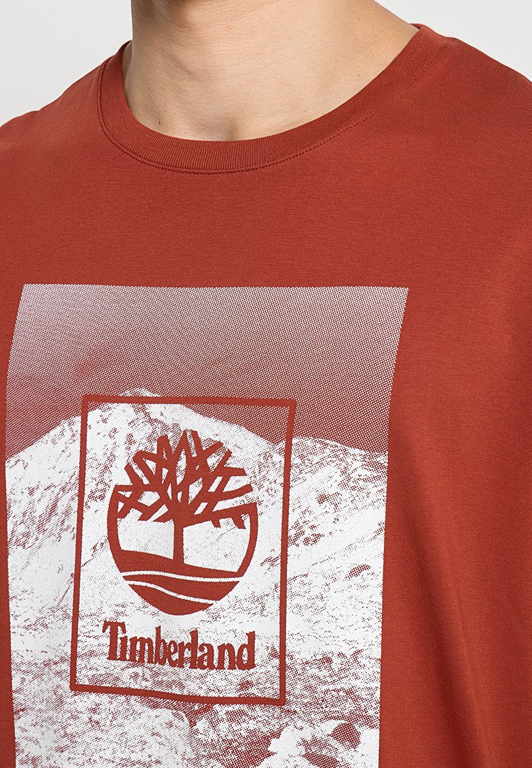 T-shirt in cotone rosso con una grafica bianca di un logo ad albero e montagne all'interno di un riquadro rettangolare, design con scollatura a girocollo e una texture liscia.