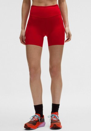 Kvinde iført lyse røde træningsshorts med høj talje, sorte sokker og røde løbesko med lyseblå snørebånd, stående med benene let adskilt.