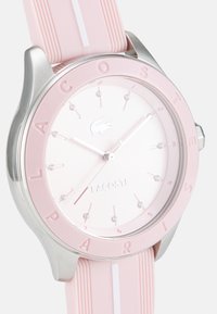 Montre Lacoste avec un bracelet en silicone rose, un boîtier en argent, un cadran blanc, des accents argentés et des marqueurs en cristal. Logo présent à 12 heures.