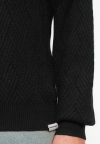 Detailný záber na rukáv a lem čierneho pleteného svetra s textúrou, s malou bielou nálepkou Cleptomanicx, nosený s svetlomodrými džínami.