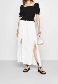 Black fitted top with puff sleeves paired with a textured white maxi skirt featuring side slits, accessorized with a light pink handbag and black sandals.