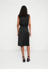 Schwarzes ärmelloses Kleid mit glatter Textur und knielangem Saum, das dezente Details aufweist. Getragen mit schwarzen, schmalen, hochhackigen Sandalen.