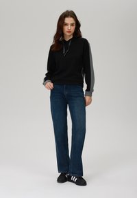 Svart hettegenser med grå ermer, kombinert med blå vid-legget jeans og svarte sneakers med hvite striper. Glatt stoffstruktur.