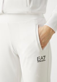Baltie sporta bikses, kas izgatavotas no gludas auduma, ar sānu kabatu un melnu EA7 Emporio Armani logo priekšpusē.