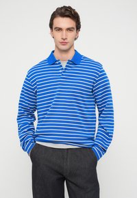 Μπλε και λευκό ριγέ polo με μακριά μανίκια, ριμπ ύφασμα στο γιακά και πόρπη με δύο κουμπιά, συνδυασμένο με σκούρο γκρι παντελόνι. Λεία υφή.