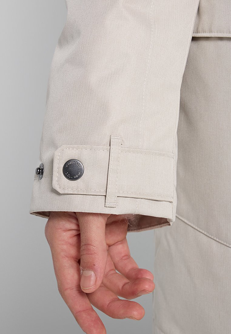 Beige Jackenärmel mit verstellbarem Riemen und metallischem Druckknopf, getragen an einer linken Hand mit entspannten und leicht gekrümmten Fingern.