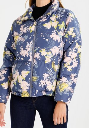 Doudoune bleue à fleurs avec des fleurs roses et jaunes, fermeture éclair sur le devant, portée sur un haut noir et un jean foncé par une personne debout.