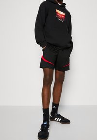 Felpa nera con cappuccio e grafica del tramonto, abbinata a pantaloni corti neri con dettagli rossi e bianchi; calze nere e scarpe adidas nere.