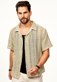 Strick-T-Shirt mit kurzen Ärmeln in hellbeige mit einem offenen Webmuster, mit einem Kragen und Vorderknöpfen versehen, über einem schwarzen Unterhemd getragen.