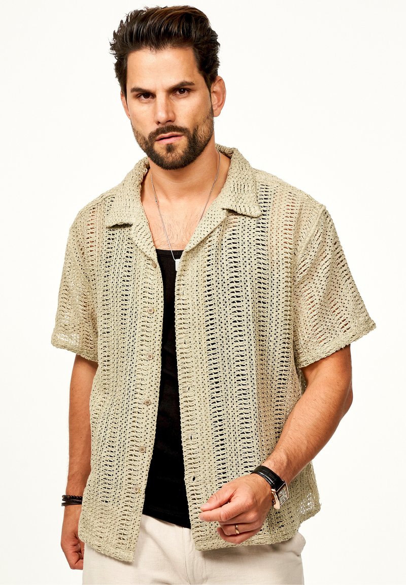 Strick-T-Shirt mit kurzen Ärmeln in hellbeige mit einem offenen Webmuster, mit einem Kragen und Vorderknöpfen versehen, über einem schwarzen Unterhemd getragen.