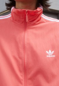 Person som bär en korallröd Adidas träningsjacka med hög krage och dragkedja samt vitt logotyp och ränder på axlarna.