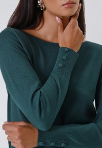 Vrouw in een donkergroen langemouwen top met stof bedekte knopen op de manchetten en zilveren abstracte oorringen, hand aan de kin.