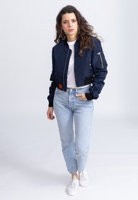 Giubbotto bomber navy con zip frontale, polsini a coste e etichetta arancione, abbinato a jeans azzurri chiari e sneakers bianche.