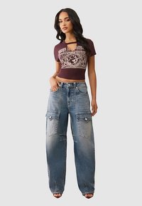 Bordowy krótki top z nadrukiem graficznym, zestawiony z luźnymi, jasnoniebieskimi jeansowymi spodniami typu cargo z kieszeniami naszywanymi i postarzanym wykończeniem.