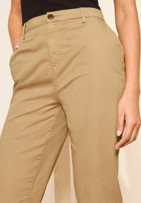 Pantaloni kaki con una superficie liscia, tasche laterali, chiusura con bottone e vestibilità rilassata, presentati su uno sfondo neutro.