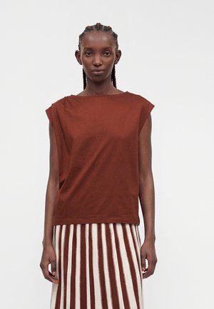 Jeune femme aux cheveux tressés portant un haut marron sans manches et une jupe à rayures verticales marron et blanches, debout devant un fond uni.