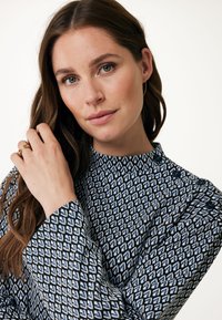 Långärmad blus i marinblått och ljusblått geometriskt mönster, med hög halsringning och knapprader vid axeln. Slät tygstruktur.