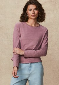 Długi rękaw, top z okrągłym dekoltem w jasnym mauve o ryflowanej fakturze z krótkim dołem. Połączony z jasnoniebieskimi dżinsami. Prosty i casualowy design.