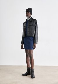 Veste courte en cuir noir avec deux grandes poches avant, portée sur un col roulé rayé, associée à une jupe en denim et des bottines noires.