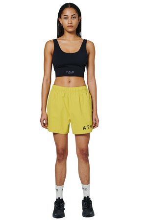 Frau steht frontal, trägt schwarzen Sport-BH, gelbe Sportshorts, weiße Socken und schwarze Turnschuhe vor weißem Hintergrund.