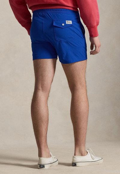 Άνδρας που φοράει μπλε σορτς με μία κουμπωτή πίσω τσέπη, κόκκινο φούτερ και λευκά αθλητικά παπούτσια, στέκεται μπροστά σε απλό μπεζ φόντο.