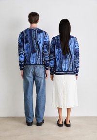 Pull en tricot bleu avec des motifs abstraits, associé à un jean bleu clair et une jupe blanche. Chaussures noires portées par les deux personnes.