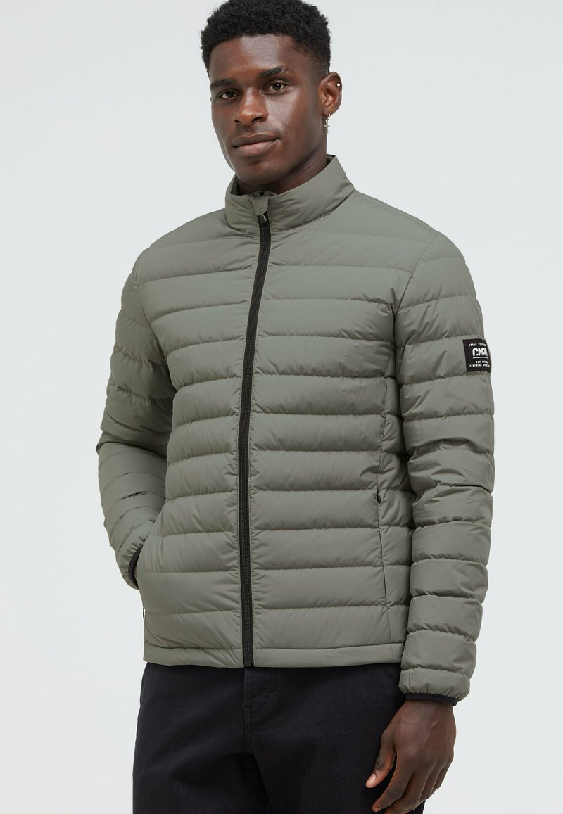Padded zip-up jacket in olive green, featuring horizontal stitching, high collar, and a logo patch on the left sleeve. Textured fabric.