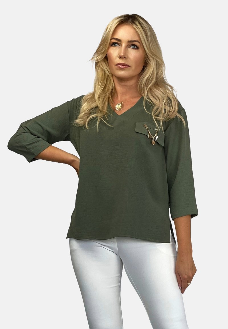 Oliwkowa bluzka z dekoltem w serek, z rękawami 3/4 i kieszonką na piersi, zdobiona ozdobnym łańcuchem z zawieszkami. Białe spodnie.