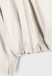 Nahaufnahme eines hellbeigen Jackenärmel und eines elastischen Saums mit sichtbaren vertikalen Nahtlinien und glatter Stofftextur.