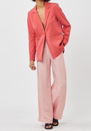 Korallenroter Blazer mit Reverskragen und zwei Vordertaschen, kombiniert mit hellrosa weit geschnittenen Hosen und schwarzen Sandalen. Glatte Stoffstruktur.
